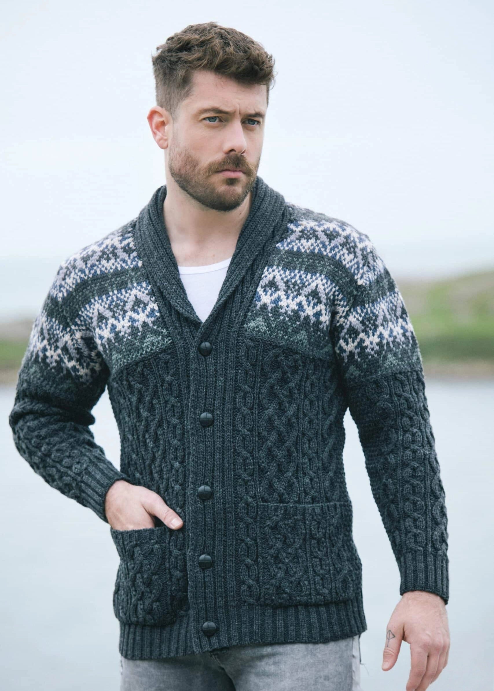 Man wearing a patterned cardigan standing outdoors with a body of water in the background