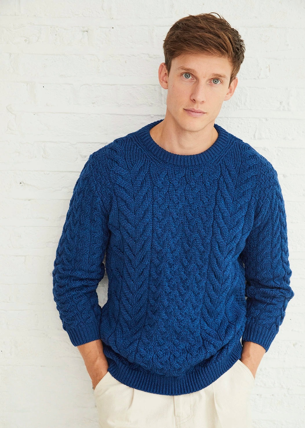 Man wearing a blue cable knit aran sweater against a white brick wall