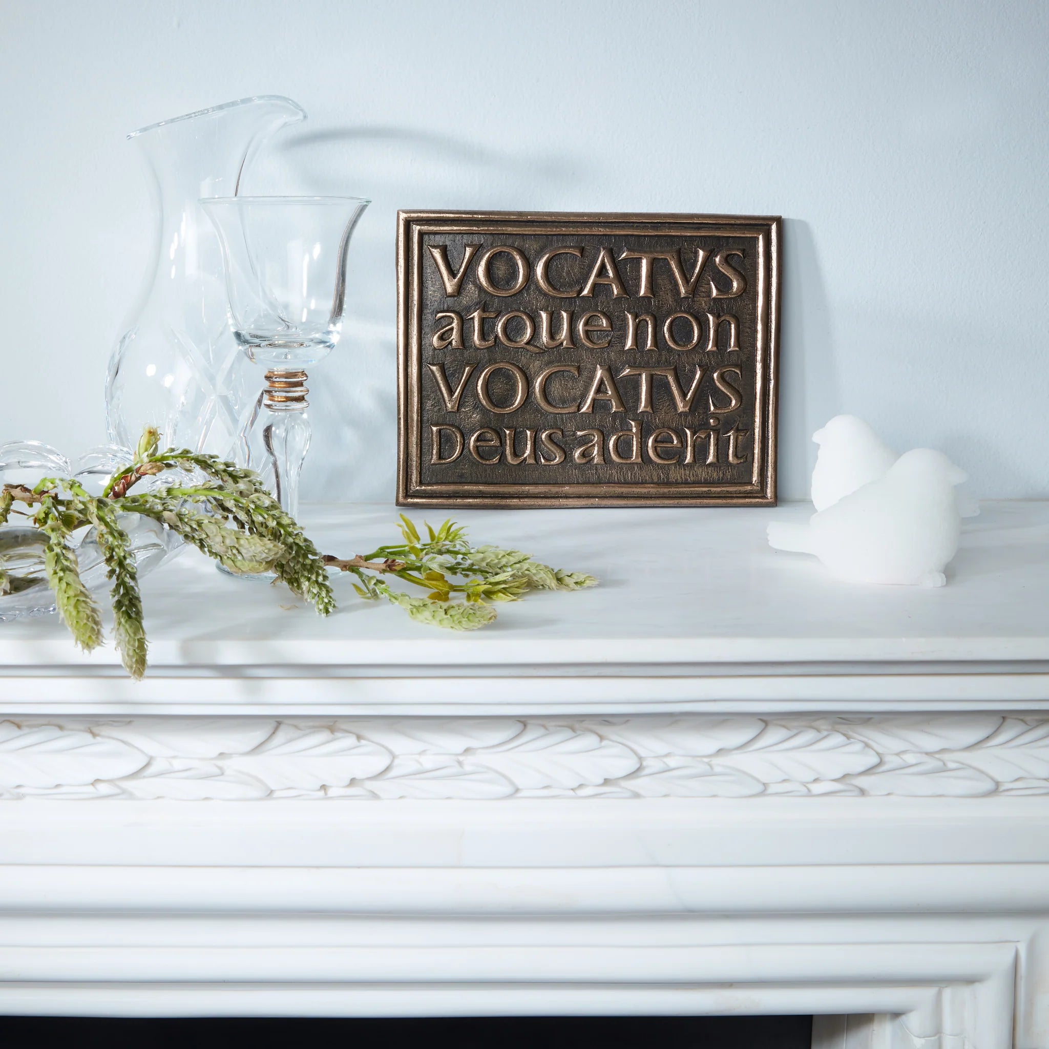 Decorative sign with Latin text on a white surface with a glass pitcher and greenery.