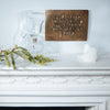 Decorative wooden plaque with engraved text on a white surface with glass pitcher and candle.