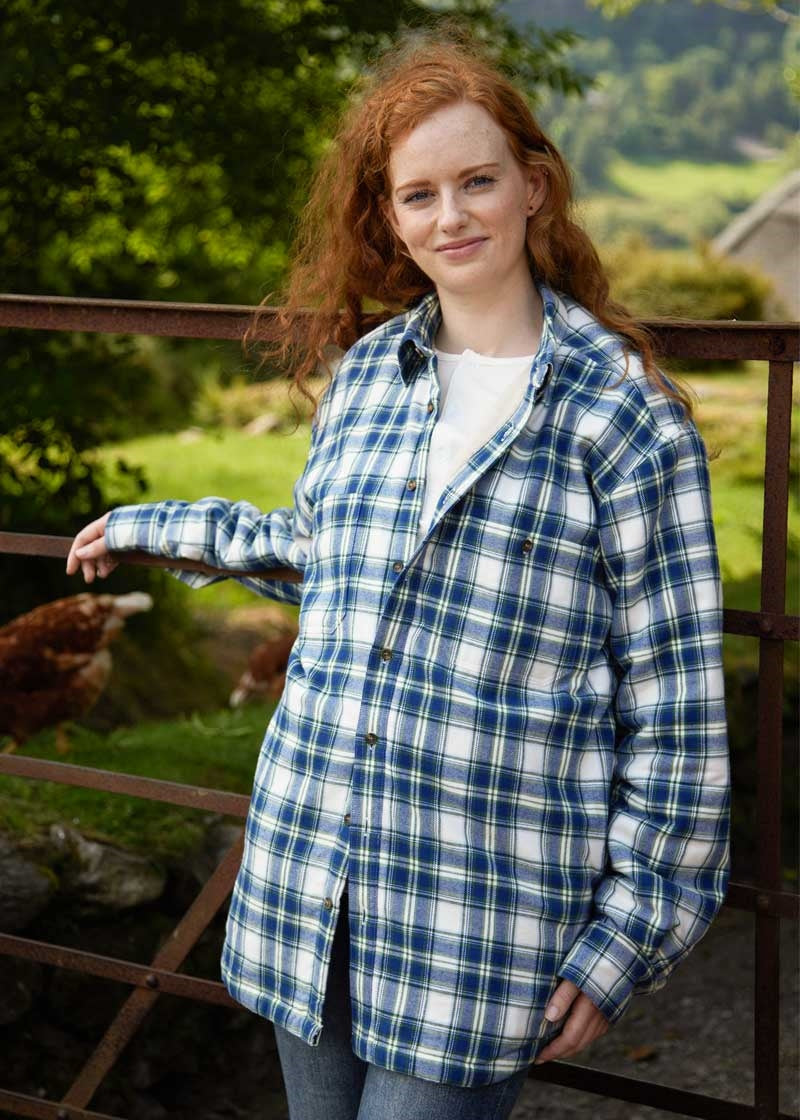 Woman wearing a blue and white checkered shirt standing outdoors with greenery in the background