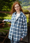 Woman wearing a blue and white checkered shirt standing outdoors with greenery in the background