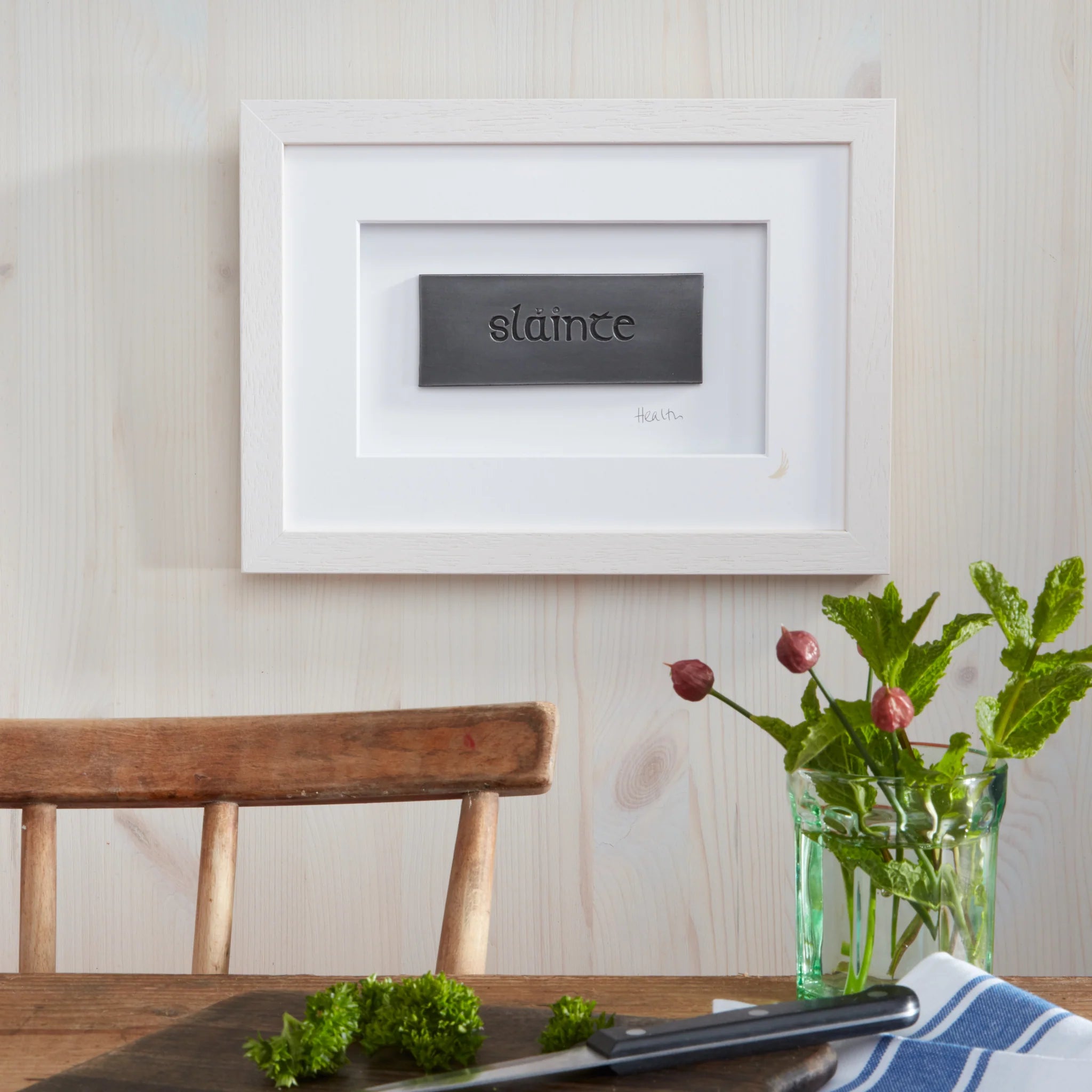 Framed artwork with 'slainte' on a wall above a wooden table with a vase of flowers.