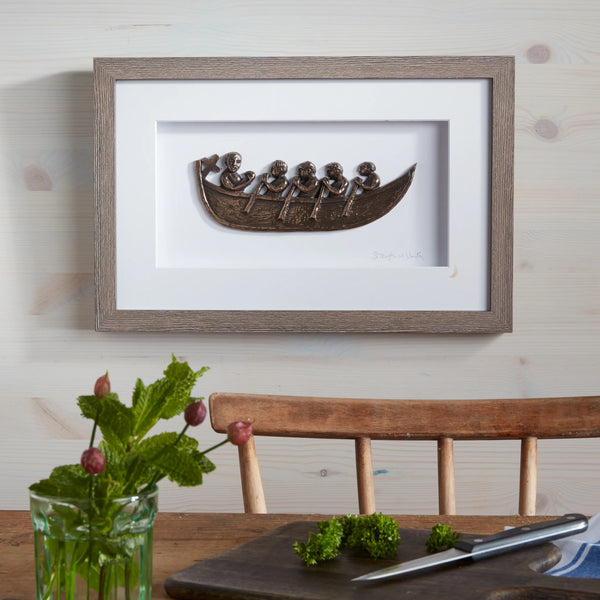 Framed artwork of a boat with figures on a wall above a wooden table.