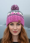 Person wearing a pink knitted beanie with a patterned design and pom-pom on a blurred natural background.