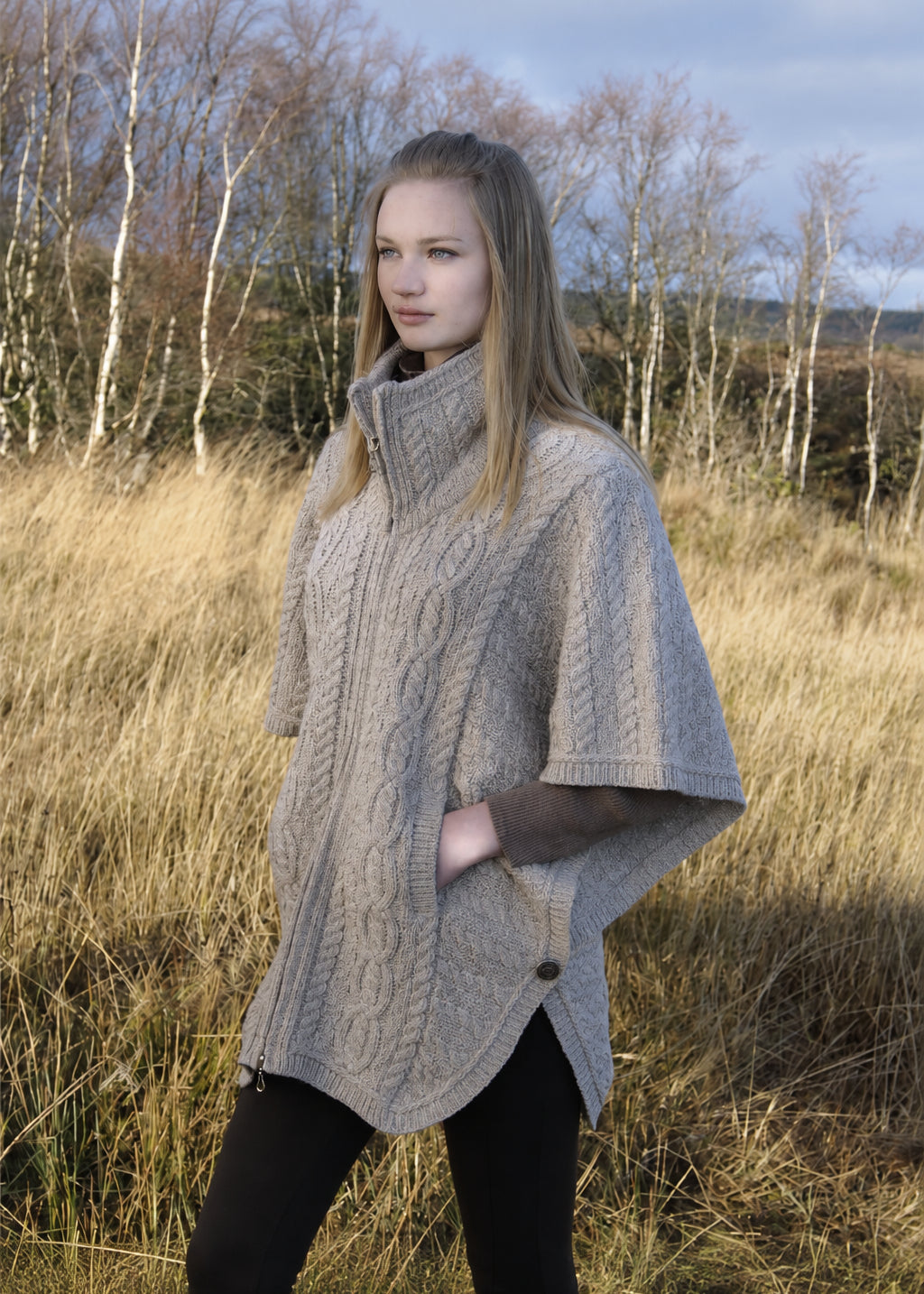 Woman wearing a knitted poncho in a natural setting with trees and grass.