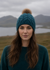 Woman wearing a blue knit beanie with a pom-pom in a natural setting with mountains and water.