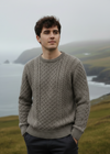 Man wearing a patterned sweater standing in a scenic landscape with water and cliffs.