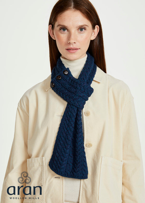 A woman wearing a cream-colored jacket and a dark navy blue cable-knit scarf, with the Aran Woolen Mills logo visible in the bottom left corner.