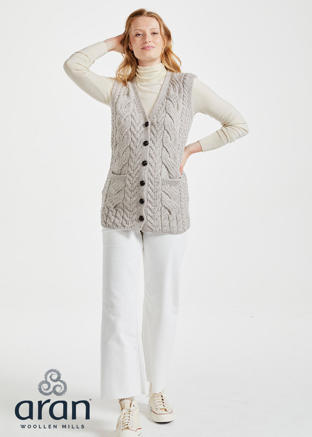 Woman wearing a cable knit vest with buttons on a white background, branded 'Aran Woollen Mills'.