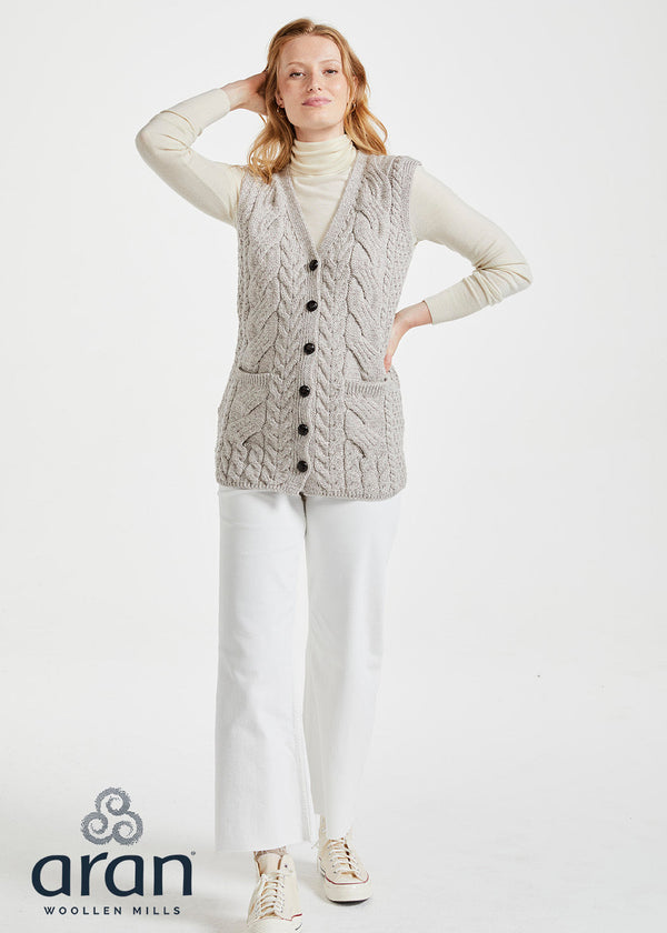 Woman wearing a cable knit vest with buttons on a white background, branded 'Aran Woollen Mills'.