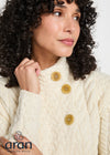 A close-up of a woman wearing a cream-colored cable-knit sweater with three large olive-green buttons. The Aran Sollen Mills brand logo is visible in the bottom-left corner.