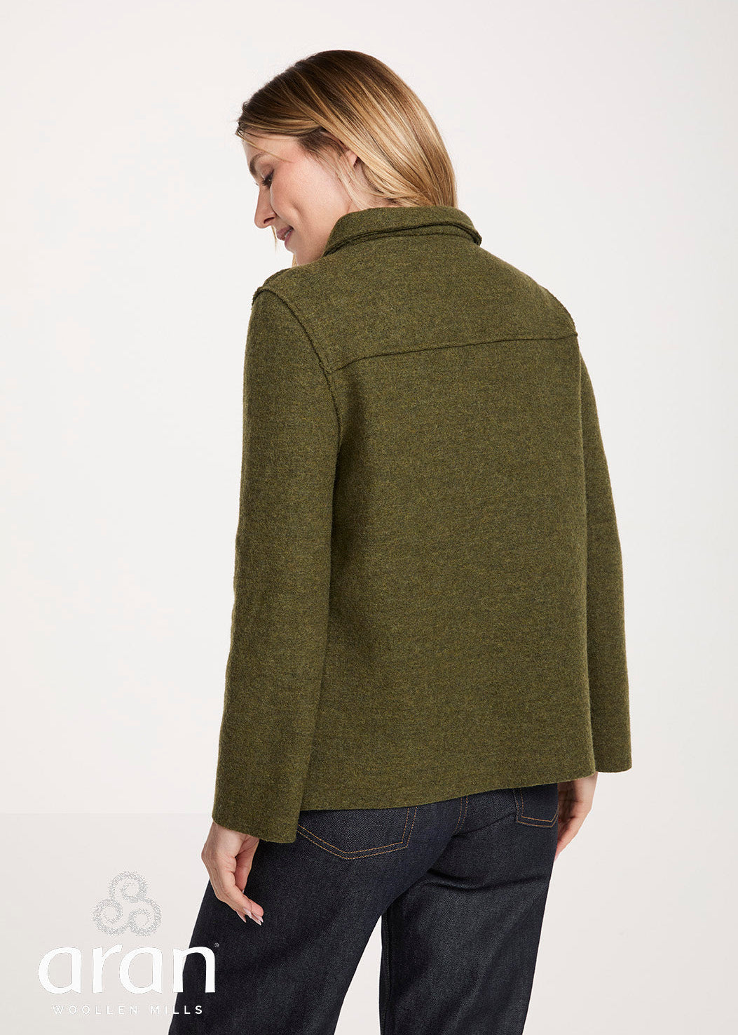 A woman shown from the back wearing an olive green wool-blend jacket and dark blue jeans, photographed against a clean white studio background. The Aran Co logo is visible in the bottom-left corner.
