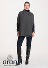 A woman modeling a dark gray cowl-neck poncho over a navy blue long-sleeve top, paired with dark blue jeans and black knee-high boots, against a light gray studio background. The Aran Woolen Mills logo is visible in the bottom-left corner.