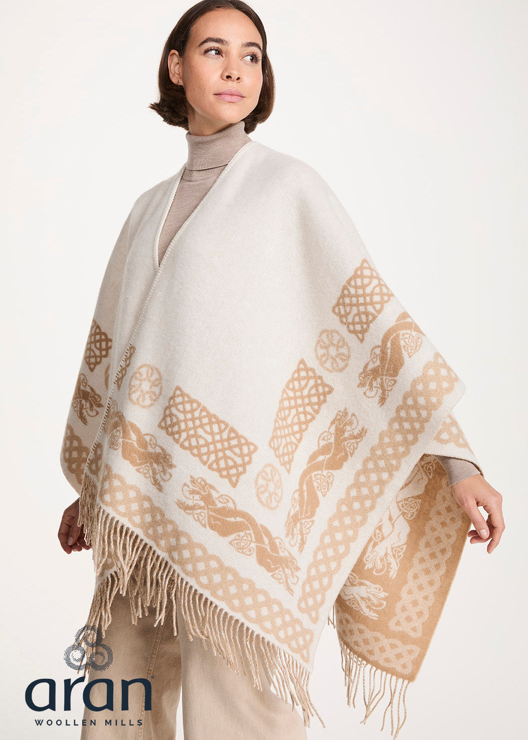 A woman wearing a cream and beige patterned Aran poncho with fringe, styled over a beige turtleneck and trousers, against a plain light background. The Aran Woolen Mills logo is visible in the bottom-left corner.