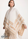 A woman wearing a cream and beige patterned Aran poncho with fringe, styled over a beige turtleneck and trousers, against a plain light background. The Aran Woolen Mills logo is visible in the bottom-left corner.