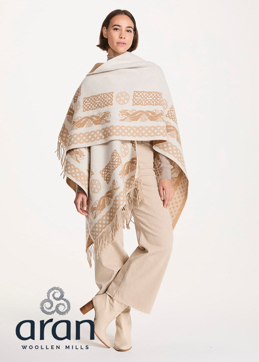 A model wearing a cream and beige patterned Aran woolen shawl with fringe, paired with beige trousers and boots, on a white studio background with the Aran Woolen Mills logo.