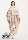 A model wearing a cream and beige patterned Aran woolen shawl with fringe, paired with beige trousers and boots, on a white studio background with the Aran Woolen Mills logo.