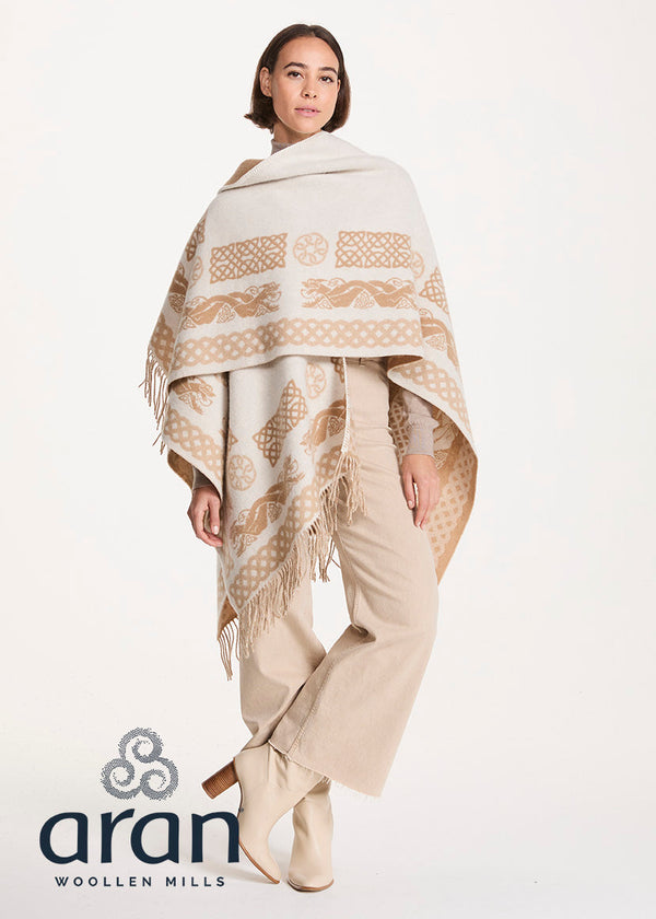 A model wearing a cream and beige patterned Aran woolen shawl with fringe, paired with beige trousers and boots, on a white studio background with the Aran Woolen Mills logo.