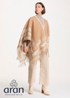 A woman modeling a beige and cream patterned Aran poncho with fringe, styled with beige trousers and cream ankle boots against a plain white studio background. The Aran Woolen Mills logo is visible in the bottom-left corner.