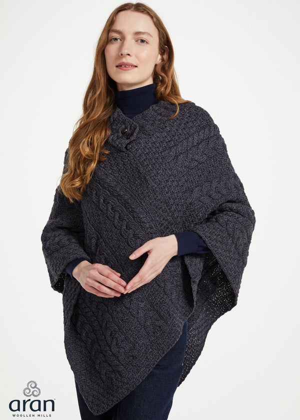 A woman wearing a dark charcoal cable-knit poncho with a large decorative flower detail on the shoulder, styled over a navy turtleneck sweater. The Aran Woolen Mills logo is visible in the bottom-left corner.