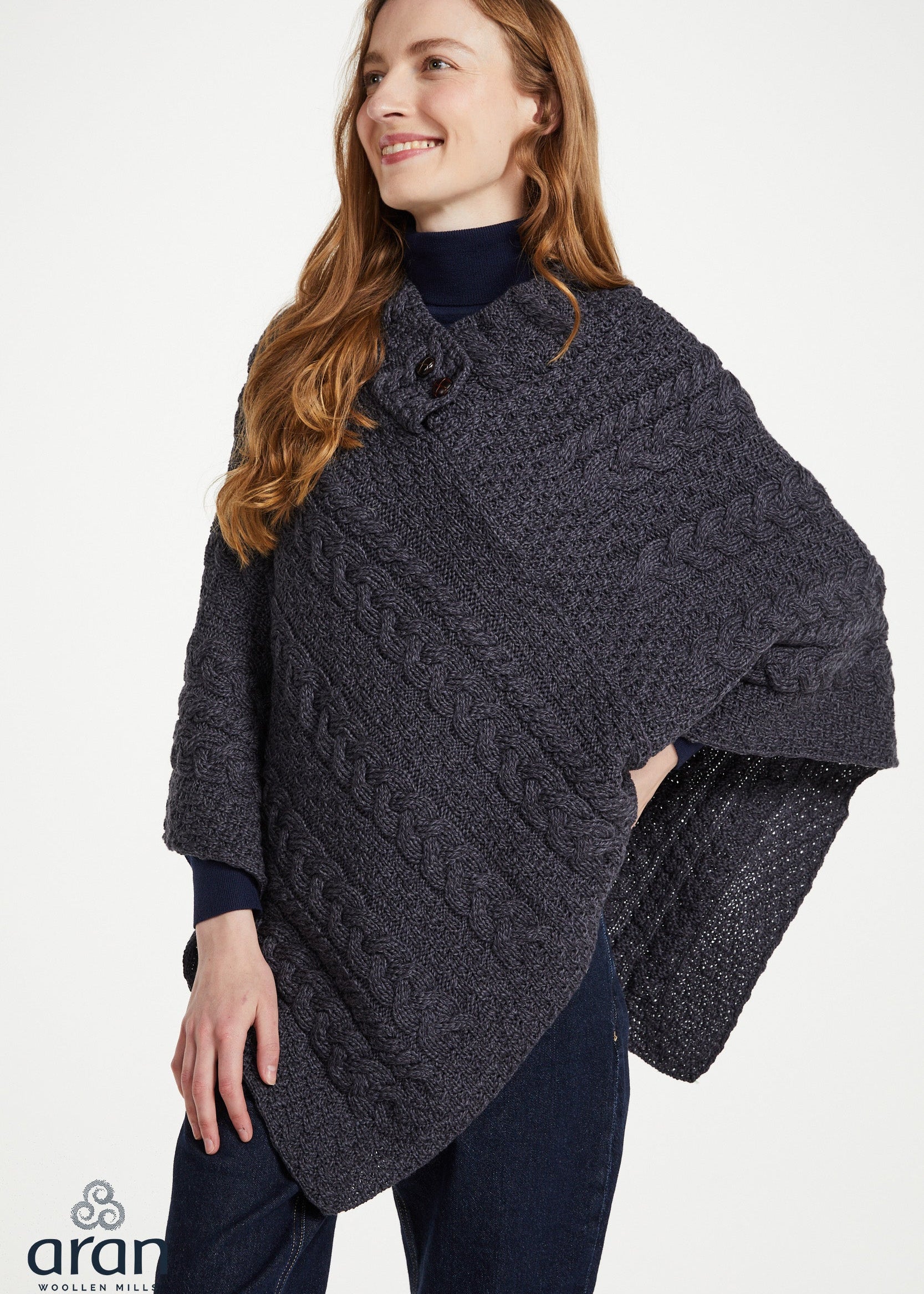A woman wearing a dark charcoal gray cable-knit poncho over a navy blue turtleneck and navy pants, photographed against a clean white studio background. The Aran logo is visible in the bottom-left corner.