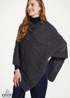 A woman wearing a dark charcoal gray cable-knit poncho over a navy blue turtleneck and navy pants, photographed against a clean white studio background. The Aran logo is visible in the bottom-left corner.
