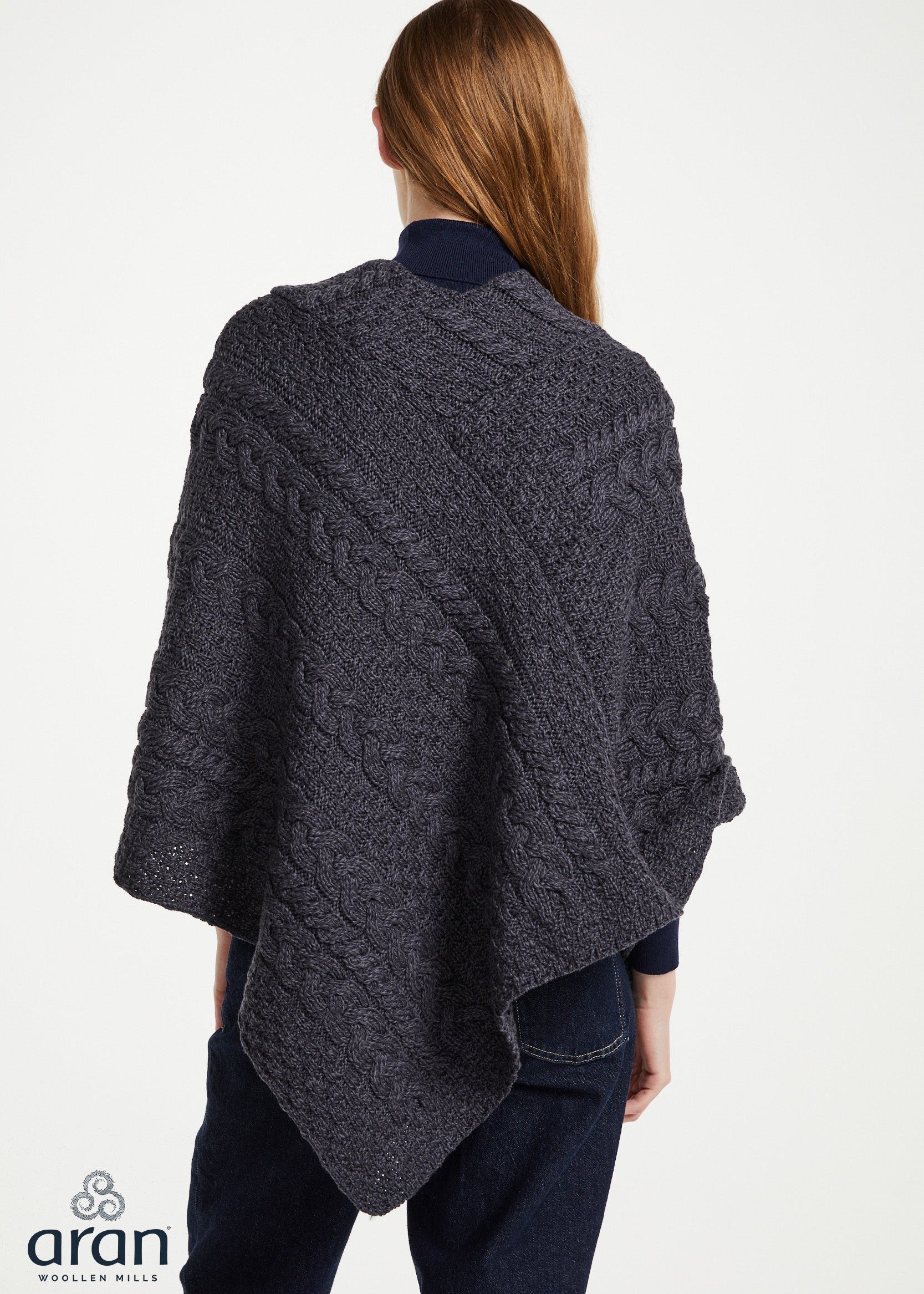 A woman shown from the back wearing a dark charcoal gray cable-knit poncho over a navy top and dark jeans, against a plain white studio background. The Aran logo is visible in the bottom-left corner.