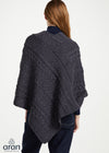 A woman shown from the back wearing a dark charcoal gray cable-knit poncho over a navy top and dark jeans, against a plain white studio background. The Aran logo is visible in the bottom-left corner.