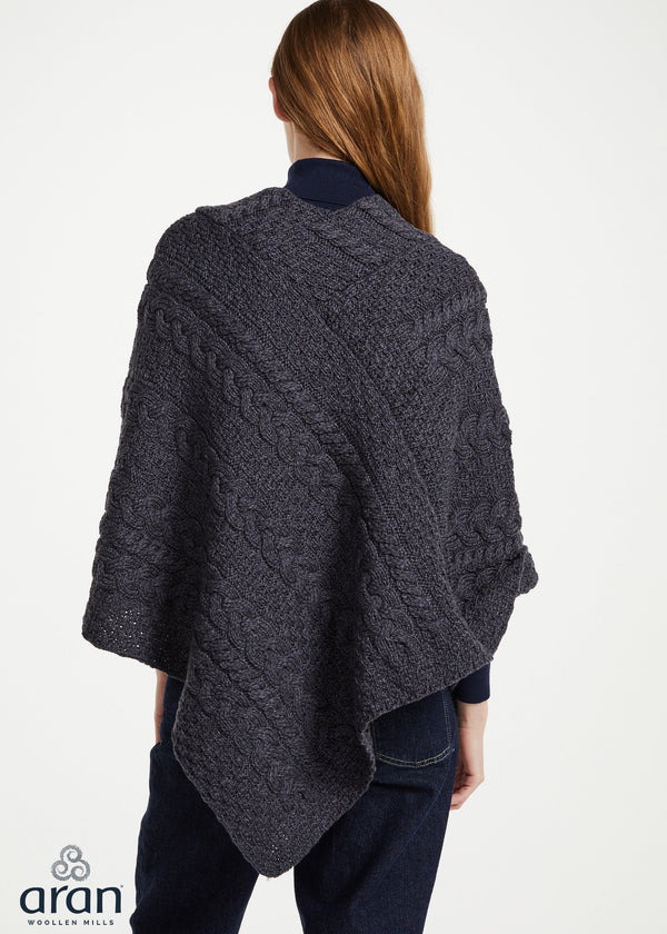 A woman shown from the back wearing a dark charcoal gray cable-knit poncho over a navy top and dark jeans, against a plain white studio background. The Aran logo is visible in the bottom-left corner.