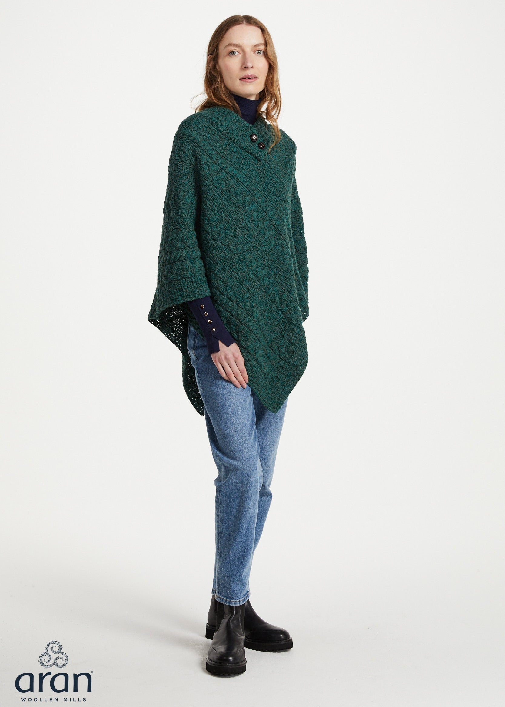 A woman modeling a dark green textured knit poncho sweater over a dark blue turtleneck, paired with blue jeans and black ankle boots, against a plain white studio background. The 'aran' brand logo is visible in the bottom-left corner.