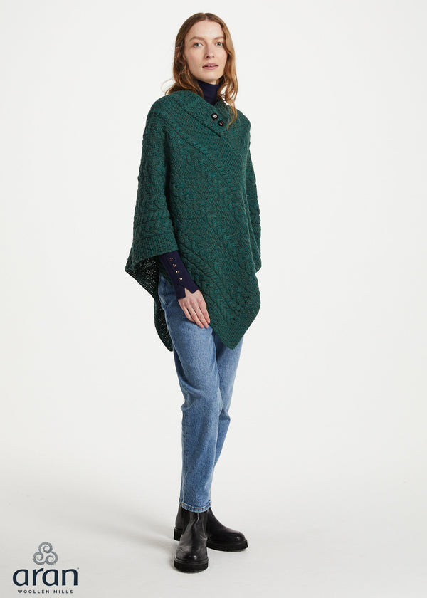 A woman modeling a dark green textured knit poncho sweater over a dark blue turtleneck, paired with blue jeans and black ankle boots, against a plain white studio background. The 'aran' brand logo is visible in the bottom-left corner.