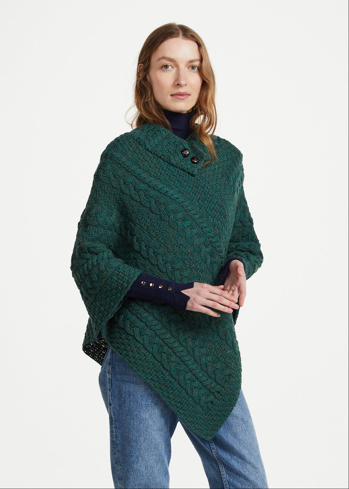 A woman modeling a dark green cable-knit poncho sweater with a high collar and asymmetrical hem, styled with blue jeans and black boots against a light gray studio background. The Aran logo is visible in the bottom-left corner.