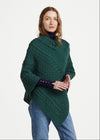 A woman modeling a dark green cable-knit poncho sweater with a high collar and asymmetrical hem, styled with blue jeans and black boots against a light gray studio background. The Aran logo is visible in the bottom-left corner.