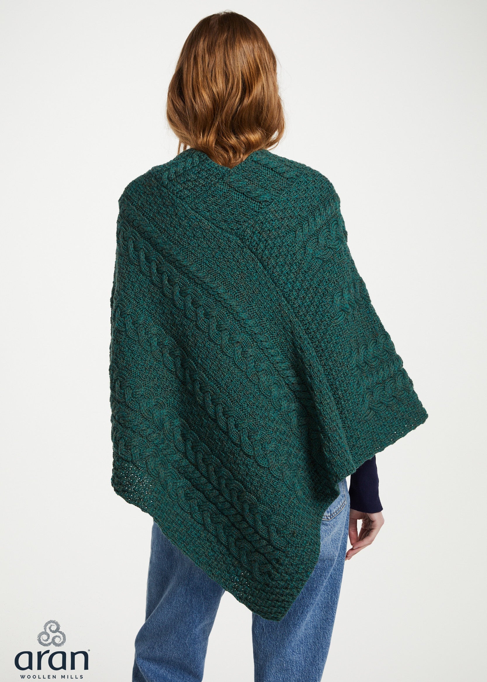 A woman shown from the back wearing a dark green cable-knit poncho or shawl over a navy top and blue jeans, against a plain light background. The Aran Weavers logo is visible in the bottom-left corner.
