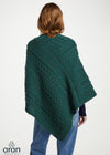 A woman shown from the back wearing a dark green cable-knit poncho or shawl over a navy top and blue jeans, against a plain light background. The Aran Weavers logo is visible in the bottom-left corner.