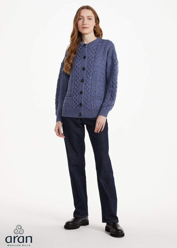 A woman modeling a blue Aran cable-knit cardigan sweater with dark navy trousers and black shoes, standing against a plain white studio background. The 'aran' brand logo is visible in the bottom-left corner.