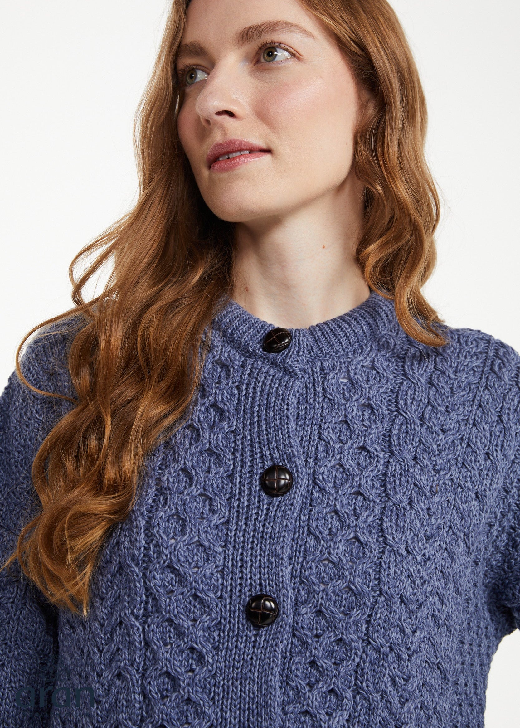 A woman with long wavy light brown hair wearing a textured blue cable-knit cardigan sweater with dark buttons, photographed against a clean white background.