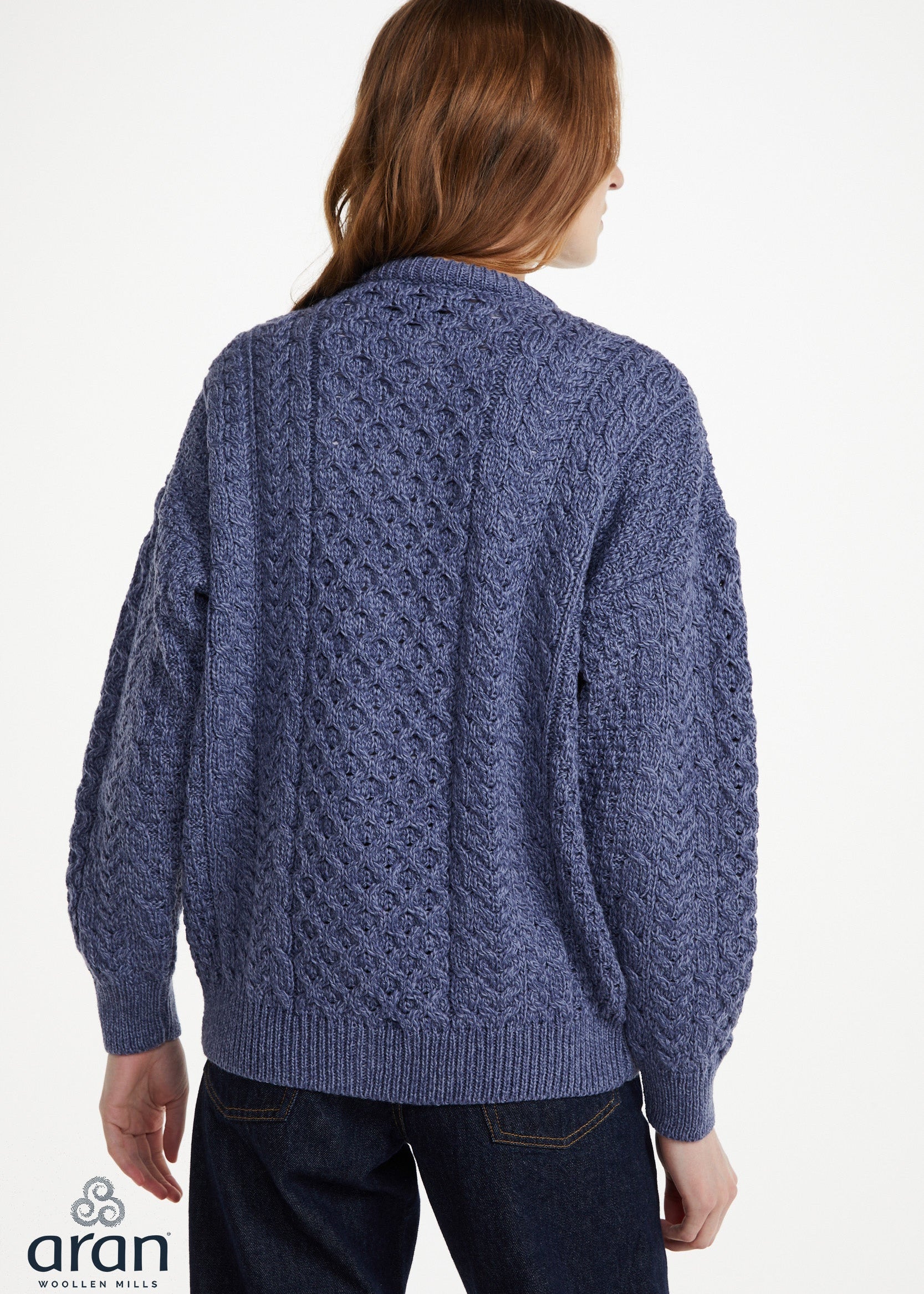 A woman viewed from the back wearing a blue cable-knit cardigan sweater and dark jeans against a plain white studio background. The Aran logo appears in the lower-left corner.