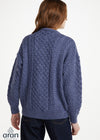 A woman viewed from the back wearing a blue cable-knit cardigan sweater and dark jeans against a plain white studio background. The Aran logo appears in the lower-left corner.