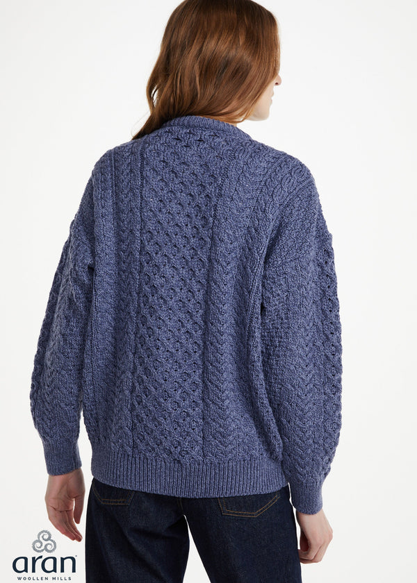 A woman viewed from the back wearing a blue cable-knit cardigan sweater and dark jeans against a plain white studio background. The Aran logo appears in the lower-left corner.