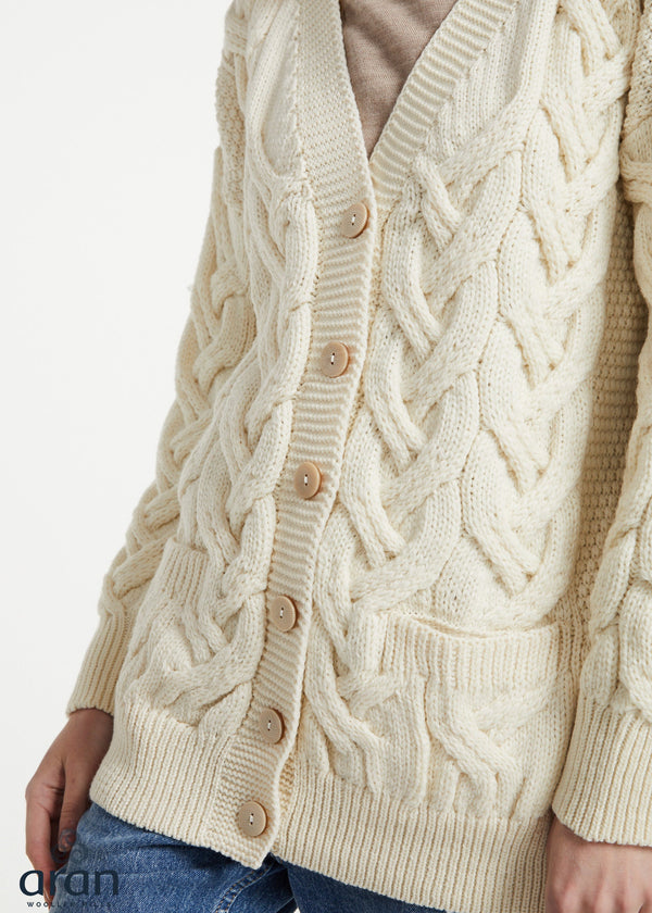 Cream cable-knit cardigan sweater with wooden buttons, worn by a model with blue jeans against a plain white background.