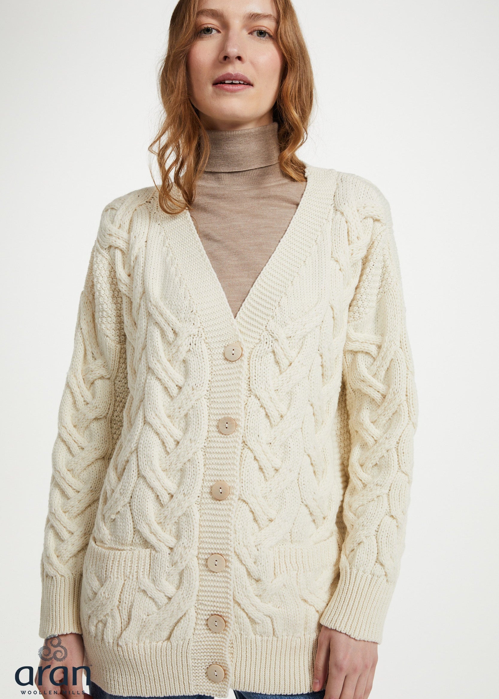 A woman wearing a cream cable-knit cardigan over a beige turtleneck sweater and blue jeans, photographed against a plain light background.