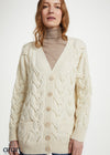 A woman wearing a cream cable-knit cardigan over a beige turtleneck sweater and blue jeans, photographed against a plain light background.