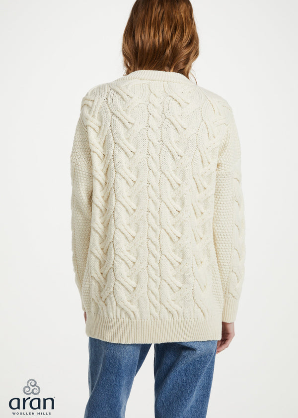 A woman viewed from the back wearing a cream cable-knit cardigan sweater and blue jeans against a plain white studio background. The Aran logo appears in the bottom-left corner.