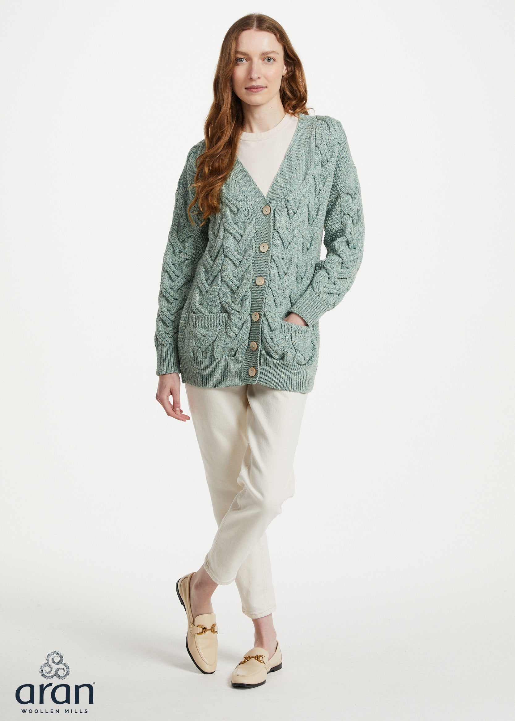 A woman modeling a sage green cable-knit cardigan with button-front closure and front pockets, paired with cream trousers and beige loafers, on a light gray studio background. The Aran logo appears in the lower-left corner.