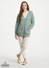 A woman modeling a sage green cable-knit cardigan with button-front closure and front pockets, paired with cream trousers and beige loafers, on a light gray studio background. The Aran logo appears in the lower-left corner.
