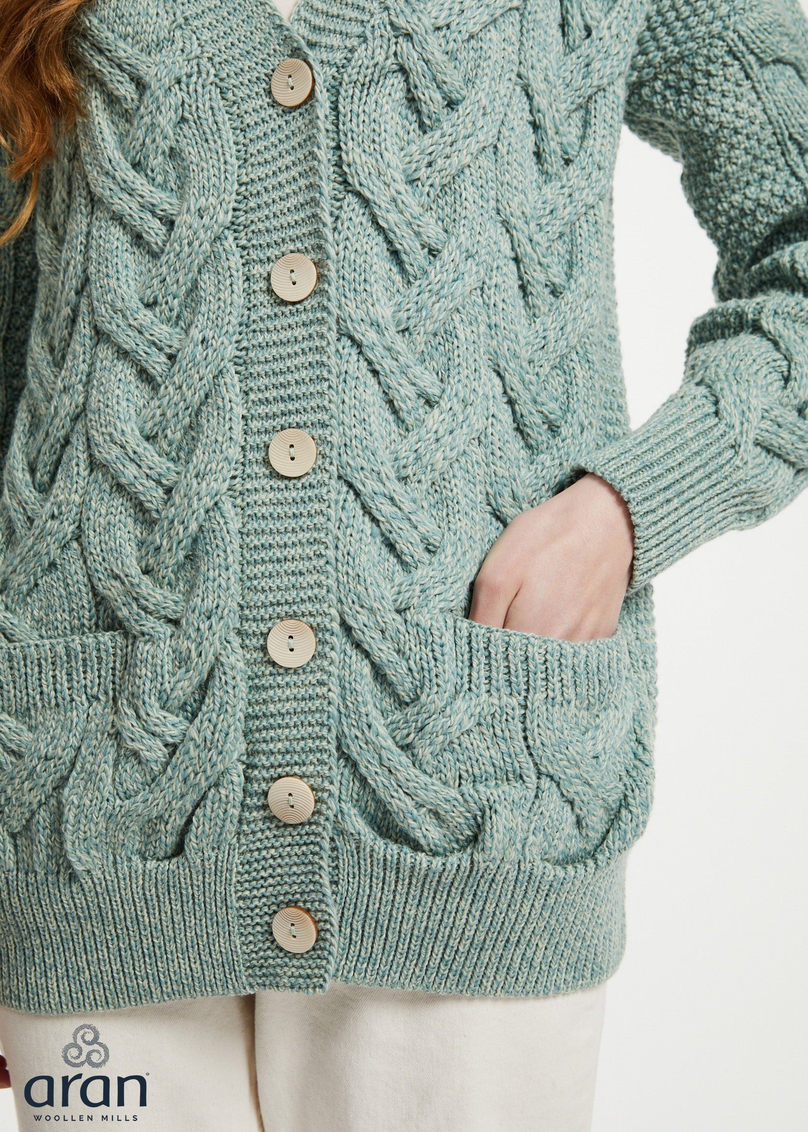 A close-up of a person wearing a sage green cable-knit cardigan sweater with wooden buttons and front pockets, paired with off-white pants. The brand logo 'aran' is visible in the bottom-left corner.