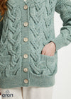 A close-up of a person wearing a sage green cable-knit cardigan sweater with wooden buttons and front pockets, paired with off-white pants. The brand logo 'aran' is visible in the bottom-left corner.