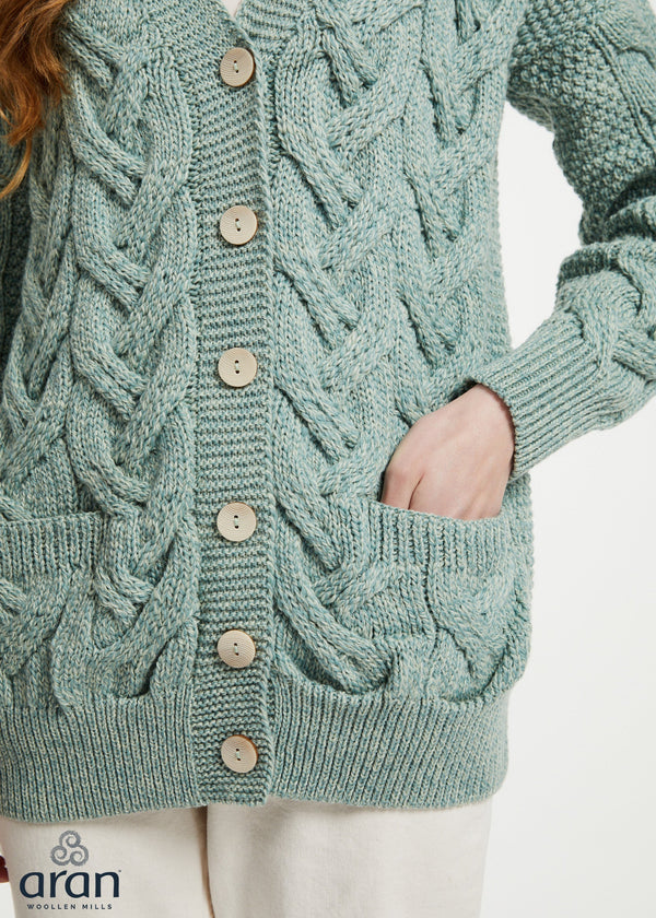 A close-up of a person wearing a sage green cable-knit cardigan sweater with wooden buttons and front pockets, paired with off-white pants. The brand logo 'aran' is visible in the bottom-left corner.